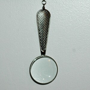 May include: A silver-toned magnifying glass pendant featuring a circular lens and a tapered handle with a textured, cross-hatched grid pattern. The piece is attached to a small metal loop at the top, designed for suspension as a necklace charm.