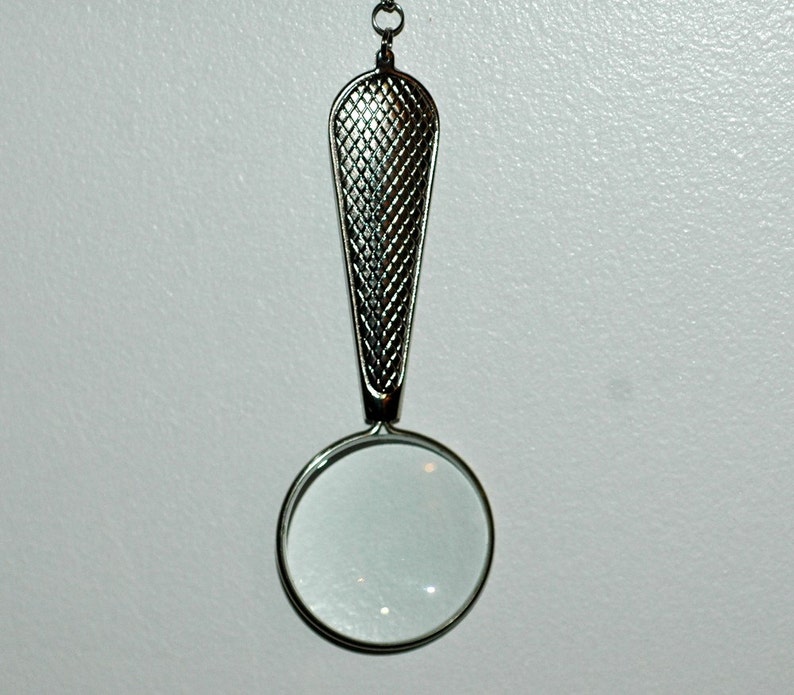 May include: A silver-toned magnifying glass pendant featuring a circular lens and a tapered handle with a textured, cross-hatched grid pattern. The piece is attached to a small metal loop at the top, designed for suspension as a necklace charm.