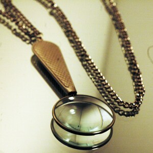 May include: A metallic pendant necklace featuring a circular magnifying glass lens encased in a bronze-toned frame. The handle of the magnifier has a textured, cross-hatched pattern and connects to a long, thin, matching metallic link chain.
