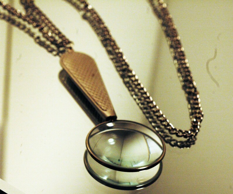 May include: A metallic pendant necklace featuring a circular magnifying glass lens encased in a bronze-toned frame. The handle of the magnifier has a textured, cross-hatched pattern and connects to a long, thin, matching metallic link chain.