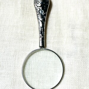 May include: A silver-toned magnifying glass pendant on a delicate chain. The handle features an ornate, embossed floral pattern with dark recessed detailing. The circular lens is framed by a thin metal rim, creating a functional jewelry piece.