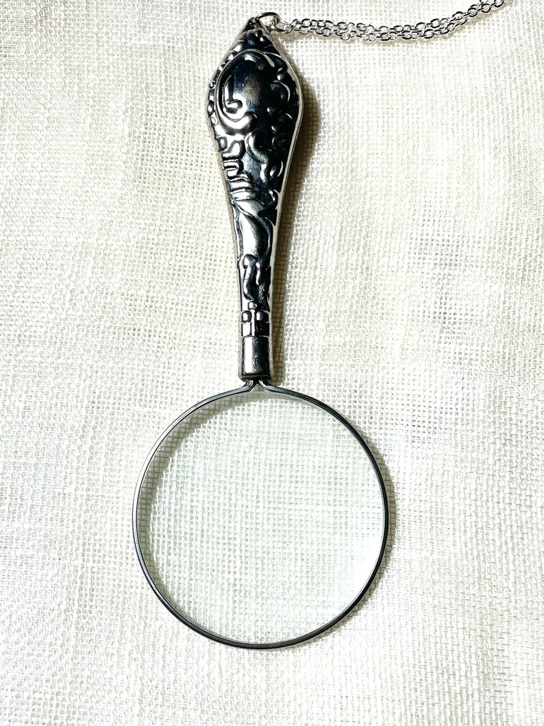 May include: A silver-toned magnifying glass pendant on a delicate chain. The handle features an ornate, embossed floral pattern with dark recessed detailing. The circular lens is framed by a thin metal rim, creating a functional jewelry piece.