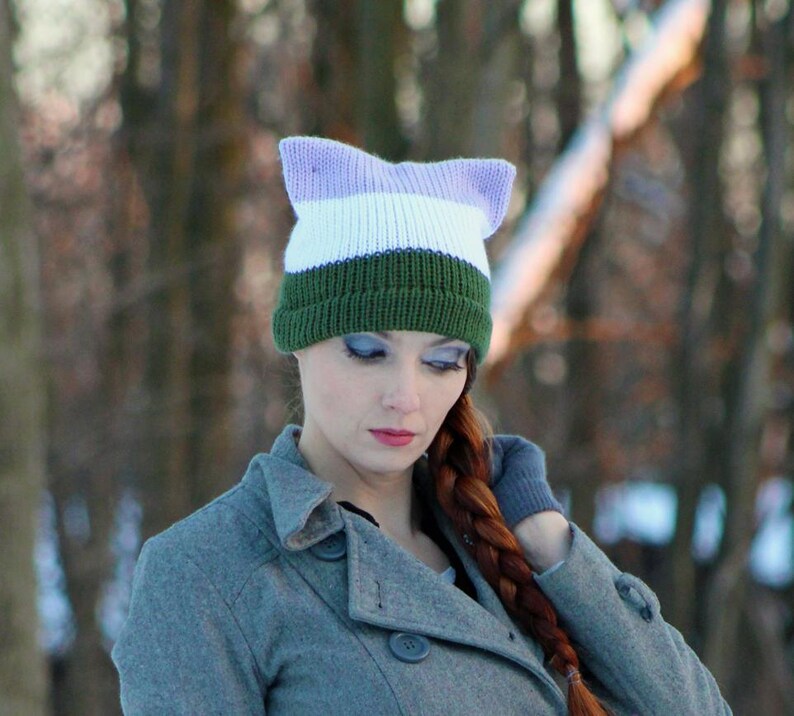 May include: A woman wearing a gray coat with a green, white, and lavender striped knit hat with cat ears.