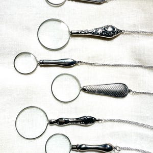 May include: Six silver-toned magnifying glass pendants, each featuring a circular lens and a unique handle design, attached to delicate silver-toned chains. The handles vary in texture, including smooth, floral embossed, and cross-hatched patterns.