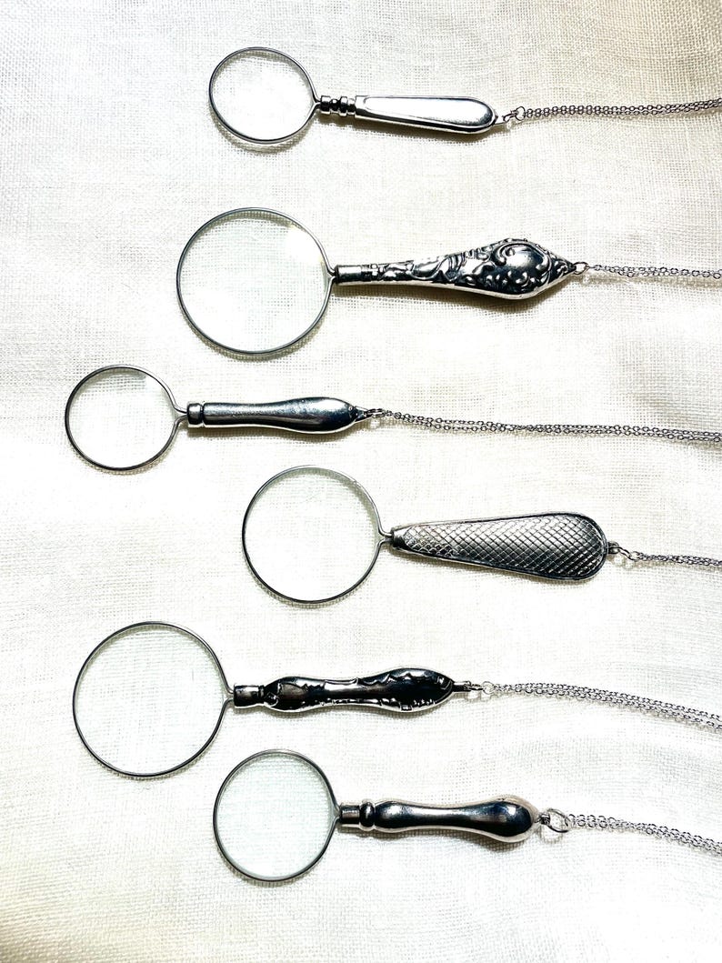 May include: Six silver-toned magnifying glass pendants, each featuring a circular lens and a unique handle design, attached to delicate silver-toned chains. The handles vary in texture, including smooth, floral embossed, and cross-hatched patterns.