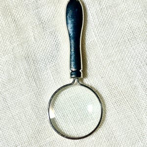 May include: A silver-toned magnifying glass pendant on a chain. The magnifying glass features a circular lens encased in a metallic frame with a smooth, elongated handle that connects to a small loop at the top for the chain attachment.
