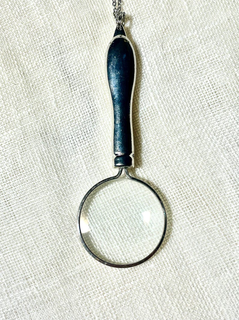 May include: A silver-toned magnifying glass pendant on a chain. The magnifying glass features a circular lens encased in a metallic frame with a smooth, elongated handle that connects to a small loop at the top for the chain attachment.