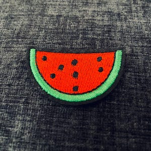 Könnte beinhalten: Ein gestickter Patch in Form einer Wassermelonen-Scheibe. Der Patch ist grün und rot mit schwarzen Kernen und einem grünen Umriss.