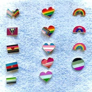 May include: A collection of 12 enamel pins featuring various pride flags and symbols, including a rainbow heart, a transgender heart, a lesbian flag, a bisexual flag, a pansexual flag, a genderqueer flag, a non-binary flag, a progress pride flag, a rainbow flag, a black and white genderqueer flag, a black and white lesbian flag, and a black and white transgender flag.