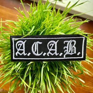 Może przedstawiać: Naszywka haftowana w kolorze czarnym i białym z tekstem "A.C.A.B." w białych literach na czarnym tle.