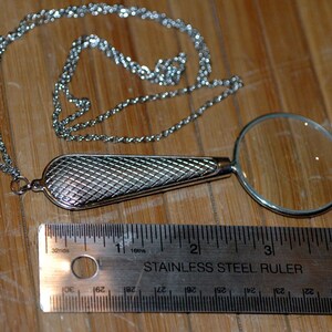 May include: A silver-toned magnifying glass pendant on a matching chain, featuring a textured, cross-hatched handle. A stainless steel ruler placed below the pendant shows the magnifying glass lens has a diameter of approximately 1.5 inches.