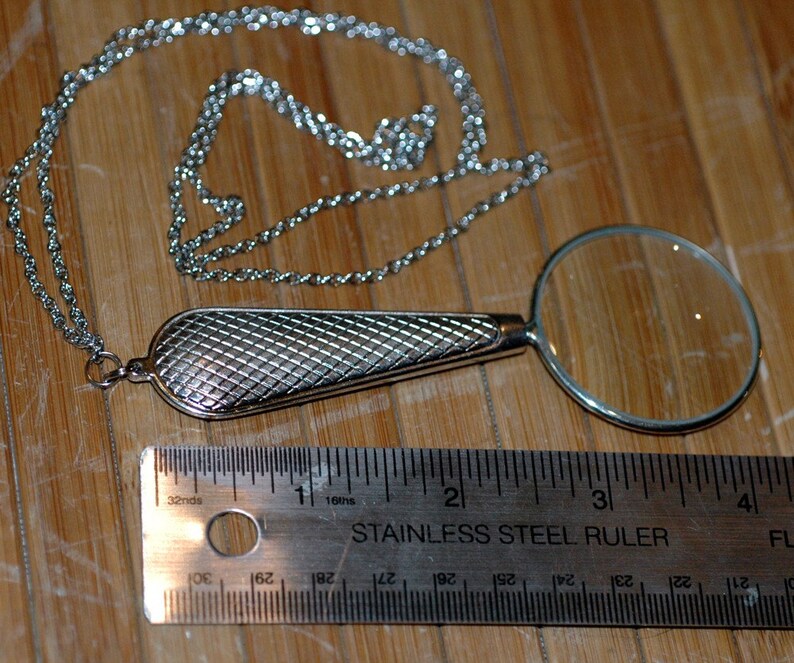 May include: A silver-toned magnifying glass pendant on a matching chain, featuring a textured, cross-hatched handle. A stainless steel ruler placed below the pendant shows the magnifying glass lens has a diameter of approximately 1.5 inches.