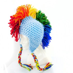 Peut inclure: Un chapeau en crochet bleu avec une crête mohawk arc-en-ciel et de longues attaches tressées. Le chapeau est sur une tête de mannequin blanche.
