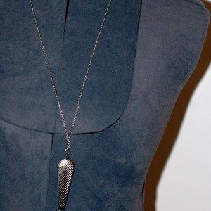 May include: A silver-toned pendant necklace featuring a long, textured, teardrop-shaped metal bail attached to a circular magnifying glass lens. The pendant hangs from a thin, matching silver-colored metal chain, creating a functional piece of jewelry.