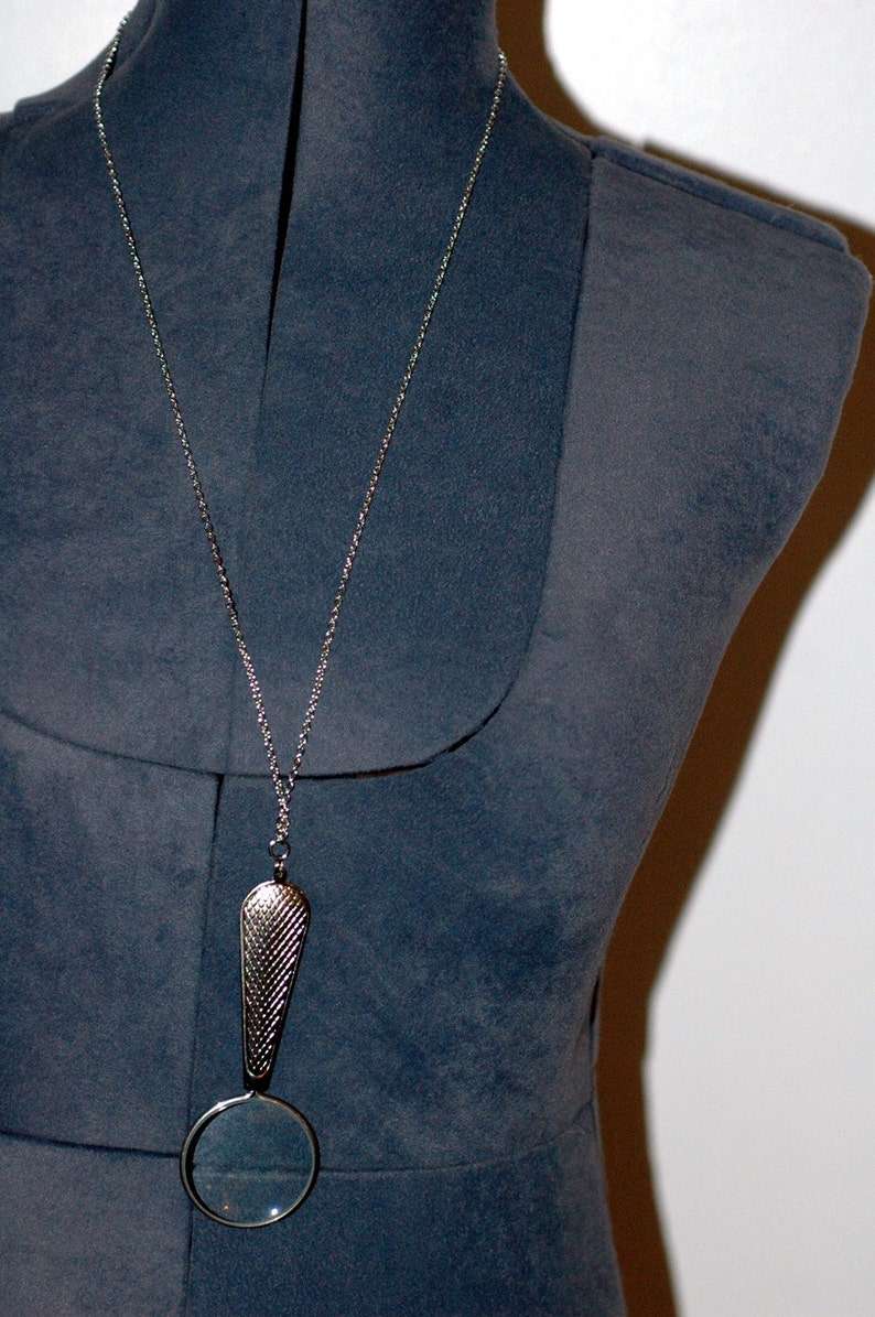 May include: A silver-toned pendant necklace featuring a long, textured, teardrop-shaped metal bail attached to a circular magnifying glass lens. The pendant hangs from a thin, matching silver-colored metal chain, creating a functional piece of jewelry.