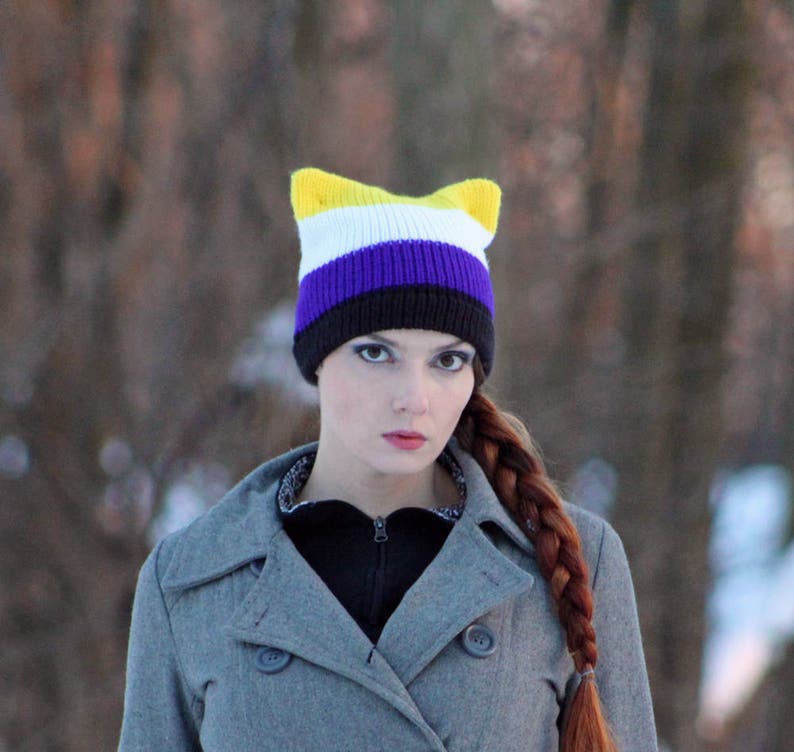 May include: A person wearing a gray coat with a yellow, white, purple, and black cat ear beanie.