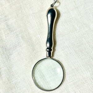 May include: A silver-toned magnifying glass pendant suspended from a delicate metal chain. The pendant features a smooth, elongated handle attached to a circular lens frame, creating a functional piece of jewelry with a polished metallic finish.