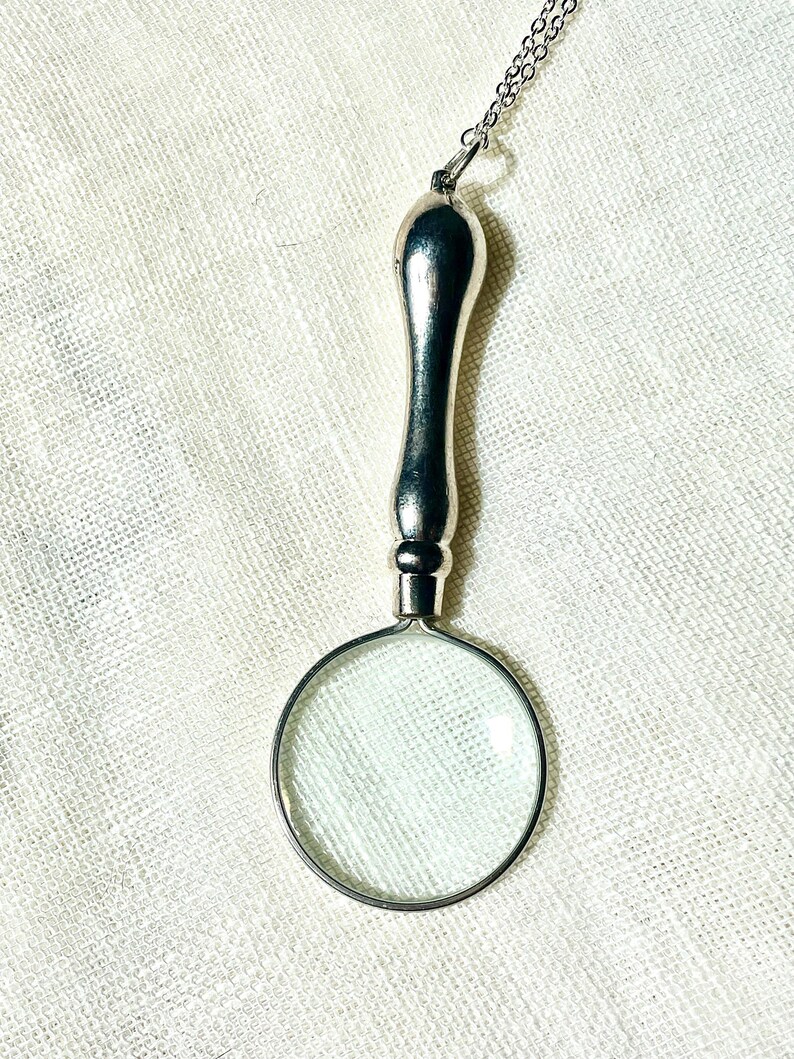 May include: A silver-toned magnifying glass pendant suspended from a delicate metal chain. The pendant features a smooth, elongated handle attached to a circular lens frame, creating a functional piece of jewelry with a polished metallic finish.