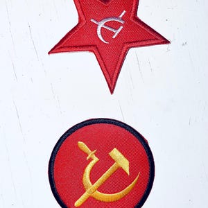 Puede incluir: Dos parches bordados, uno una estrella roja con un martillo y una hoz blancos en su interior, el otro un círculo rojo con un borde negro y un martillo y una hoz dorados en su interior.
