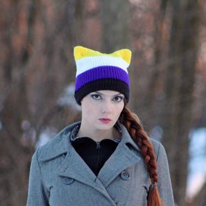 May include: A person wearing a gray coat with a yellow, white, purple, and black cat ear beanie.