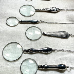 May include: Six silver-toned magnifying glass pendants arranged in a vertical row. Each pendant features a circular glass lens set in a metal frame with a unique handle design, including smooth, floral embossed, and cross-hatched textured patterns.