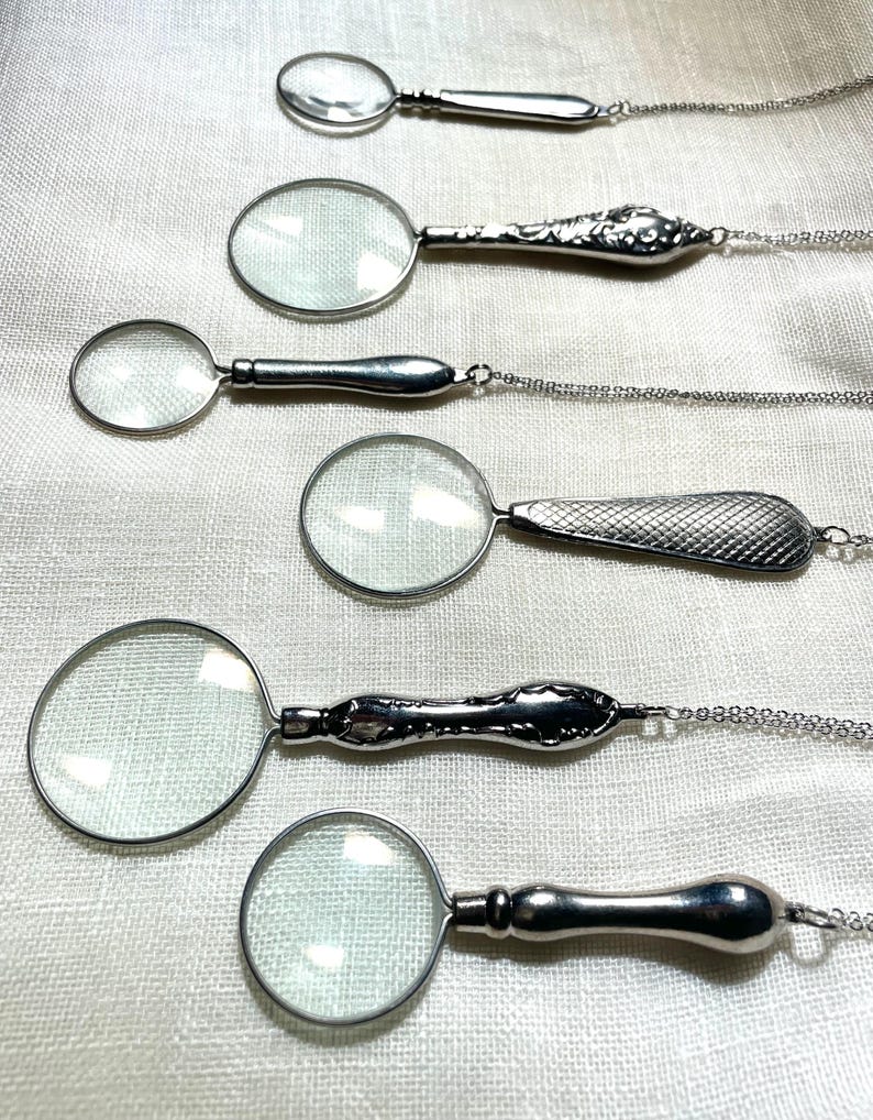 May include: Six silver-toned magnifying glass pendants arranged in a vertical row. Each pendant features a circular glass lens set in a metal frame with a unique handle design, including smooth, floral embossed, and cross-hatched textured patterns.