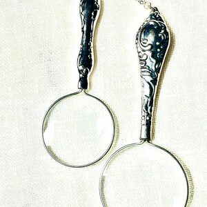 May include: Two silver-toned magnifying glass pendants on thin metal chains. Each pendant features a circular glass lens attached to a decorative, ornate handle with intricate embossed detailing. The handles resemble vintage silverware patterns.