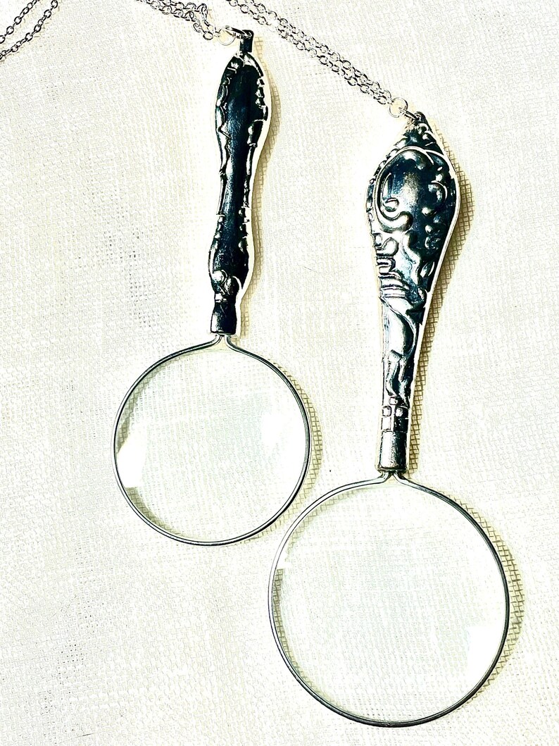 May include: Two silver-toned magnifying glass pendants on thin metal chains. Each pendant features a circular glass lens attached to a decorative, ornate handle with intricate embossed detailing. The handles resemble vintage silverware patterns.