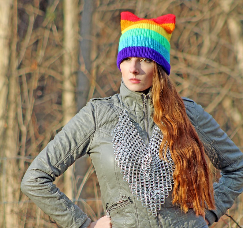 May include: A woman wearing a rainbow knit beanie with cat ears and a silver chainmail necklace. She is wearing a gray jacket and has long red hair.