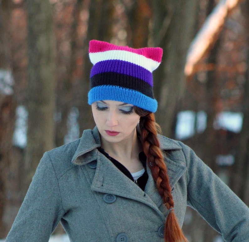 May include: A woman wearing a gray coat with a blue, black, purple, white, and pink cat ear beanie.