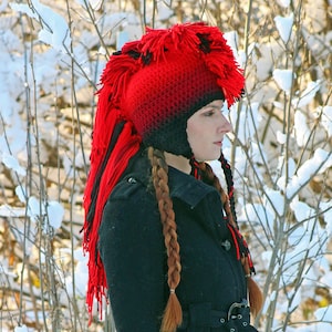 May include: A woman wearing a red and black crocheted mohawk hat with long braids. The hat has a black pom-pom on top and a black and red fringe. The woman is standing in front of a snowy background.