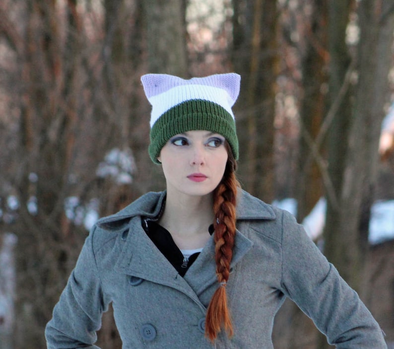 May include: A woman with long red hair braided down her back is wearing a gray coat and a green, white, and purple knit hat with cat ears.