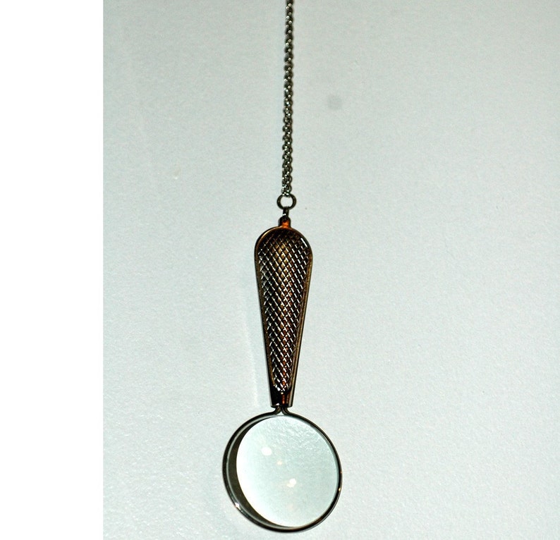 May include: A silver-toned magnifying glass pendant suspended from a delicate metal chain. The handle features a textured, cross-hatched diamond pattern, while the circular lens is framed in a smooth, polished silver-colored metal rim.