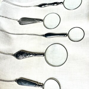 May include: Six silver-toned magnifying glass pendants on delicate matching chains. Each pendant features a circular lens with a unique metal handle design, ranging from smooth polished surfaces to intricate embossed patterns and textured cross-hatching.
