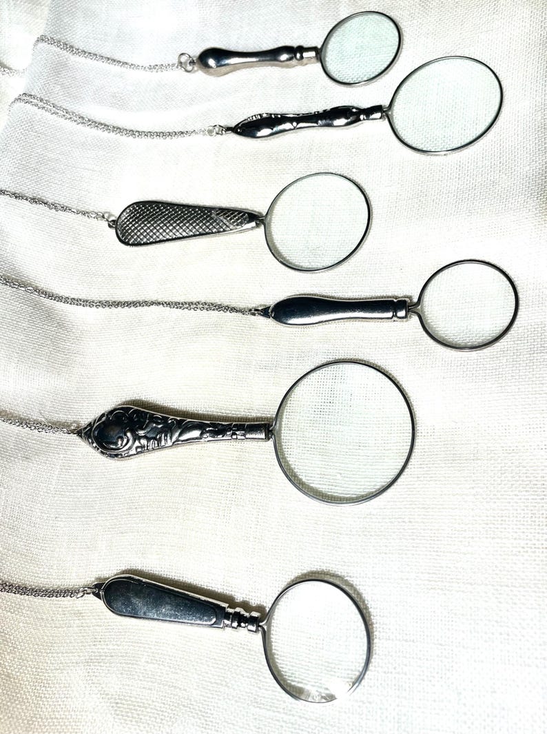 May include: Six silver-toned magnifying glass pendants on delicate matching chains. Each pendant features a circular lens with a unique metal handle design, ranging from smooth polished surfaces to intricate embossed patterns and textured cross-hatching.