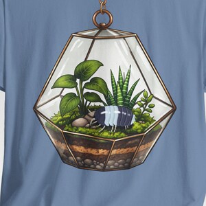May include: A light blue t-shirt featuring a detailed illustration of a terrarium. The terrarium is a geometric glass container with various plants, soil, and small decorative elements. The design is centered on the shirt.