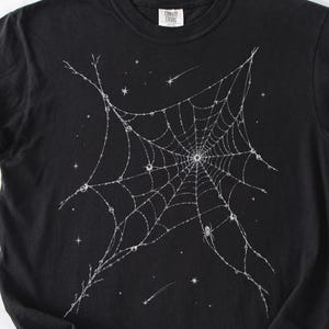 May include: A black short-sleeved t-shirt featuring a white graphic print of a spider web. The web design is embellished with small stars, crescent moons, and shooting stars scattered throughout the intricate, thin, white line-work pattern.