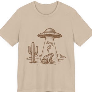 May include: Beige t-shirt featuring a brown illustration of a UFO beaming up a frog and tadpoles in a desert scene with a cactus and mountains. The design has a vintage, sketched style.