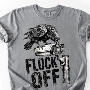 May include: A grey short-sleeve t-shirt featuring a black and white graphic of a raven perched on a security camera. The text FLOCK OFF is printed in a distressed black font below the camera, which shows sparks emitting from the lens area.