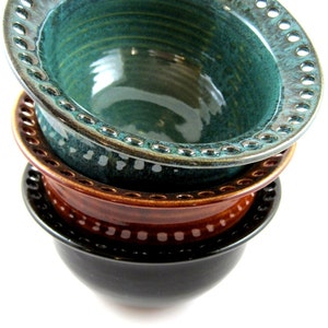 May include: Three ceramic bowls stacked on top of each other. The top bowl is teal green with a scalloped edge and a ring of holes around the rim. The middle bowl is brown with a scalloped edge. The bottom bowl is black.