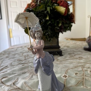 以下が含まれることがあります： ビクトリア朝時代のドレスを着た女性の磁器製フィギュアで、小さな白いパラソルを持っています。フィギュアは、水色とラベンダー色のドレス、ピンクのジャケット、お揃いの帽子が特徴です。女性の髪はカールにスタイリングされています。