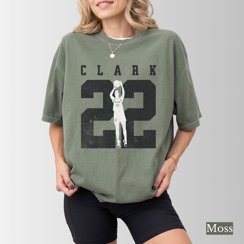 May include: Moss green t-shirt with the name "CLARK" above the number "22" in black. A silhouette of a basketball player is in the center of the number "22". The shirt is a casual style.
