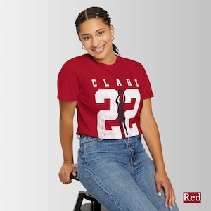 May include: Red t-shirt with the word "CLARI" above the number "22" in white, with a silhouette of a person playing basketball. The shirt is made of a soft material and is suitable for casual wear.