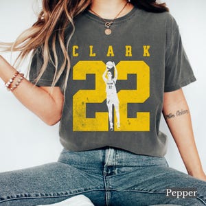 May include: Dark gray t-shirt with the name "CLARK" in yellow block letters above a large yellow number "22". A graphic of a basketball player in a white uniform is in the center of the number. The word "Pepper" is at the bottom.