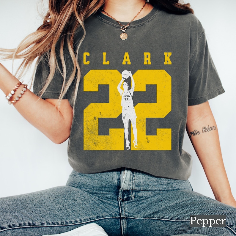 May include: Dark gray t-shirt with the name "CLARK" in yellow block letters above a large yellow number "22". A graphic of a basketball player in a white uniform is in the center of the number. The word "Pepper" is at the bottom.