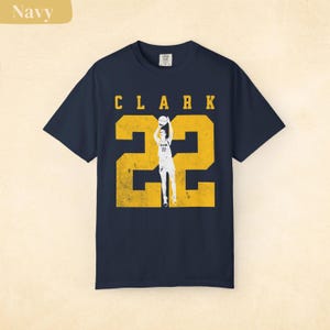 May include: Navy blue t-shirt with the name "CLARK" in yellow block letters above a large yellow number "22". A graphic of a basketball player in a shooting pose is within the number. The shirt is a classic crew neck style.