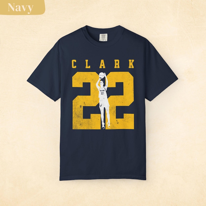May include: Navy blue t-shirt with the name "CLARK" in yellow block letters above a large yellow number "22". A graphic of a basketball player in a shooting pose is within the number. The shirt is a classic crew neck style.