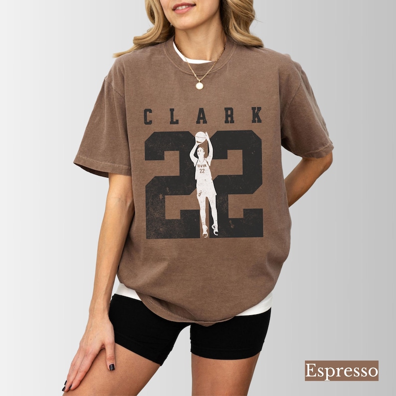 May include: Espresso-colored t-shirt featuring the name "CLARK" above the number "22" in a distressed, black font. A graphic of a basketball player is centered within the number. The shirt is a relaxed fit.