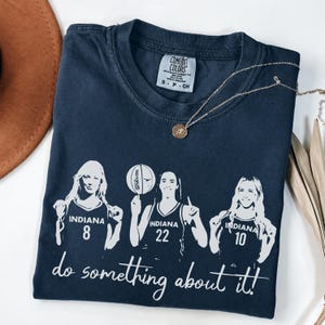 May include: Navy blue t-shirt with a graphic of three basketball players with the text "INDIANA" and numbers 8, 22, and 10. The text "do something about it!" is below the players. A gold necklace is draped across the shirt.
