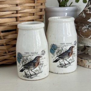 May include: Two white ceramic jars with a vintage design. Each jar features a detailed illustration of a bird, with the words "CARTE POSTALE" printed above. The jars have a slightly aged appearance, adding to their antique charm.